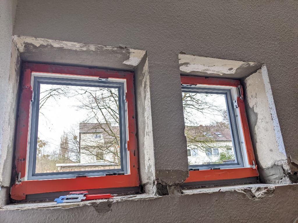 Two windows with red frames in unfinished interior; view outside onto houses and trees.