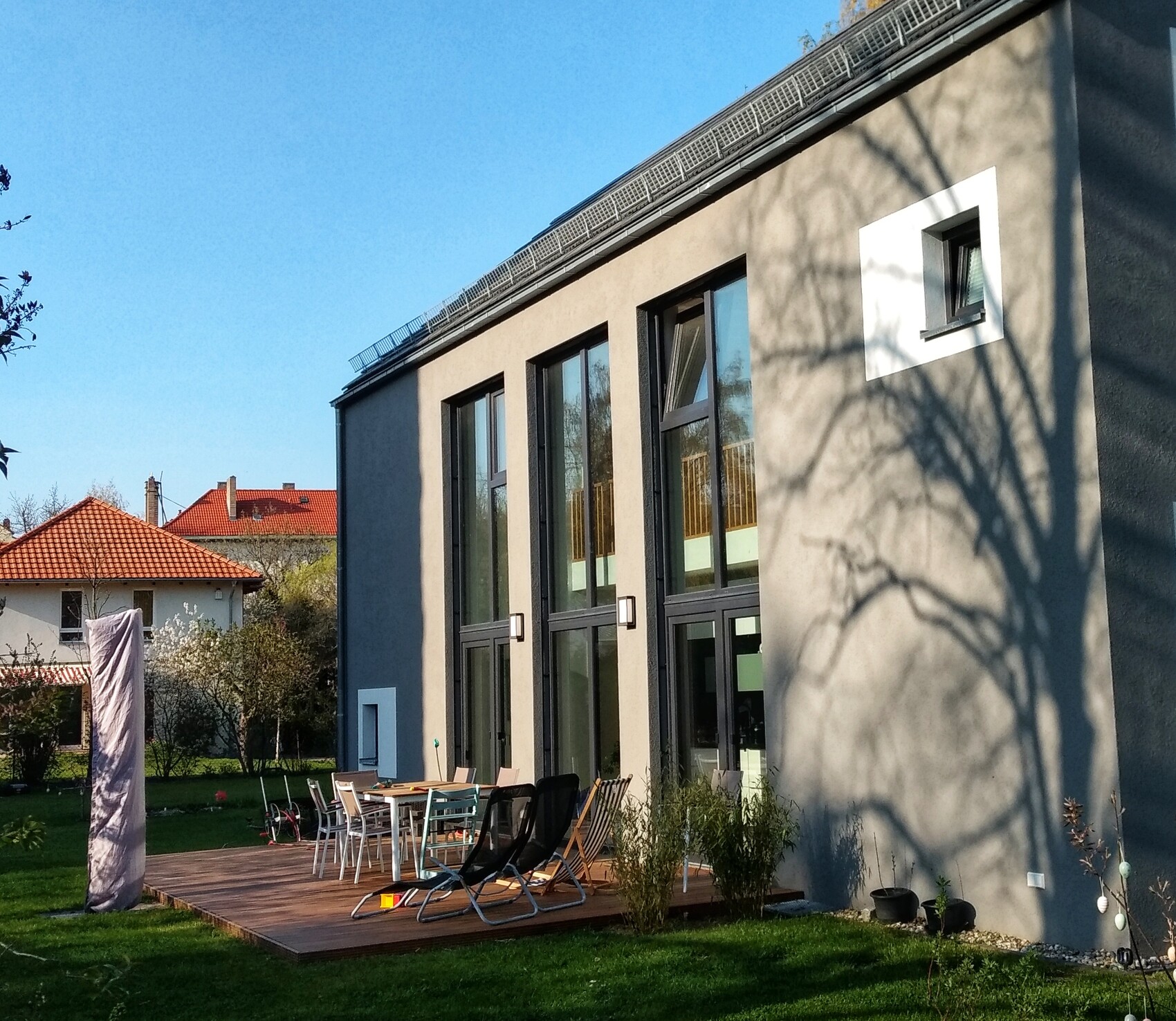 Modern building facade with large windows, wooden terrace, garden, and tree shadows on the wall