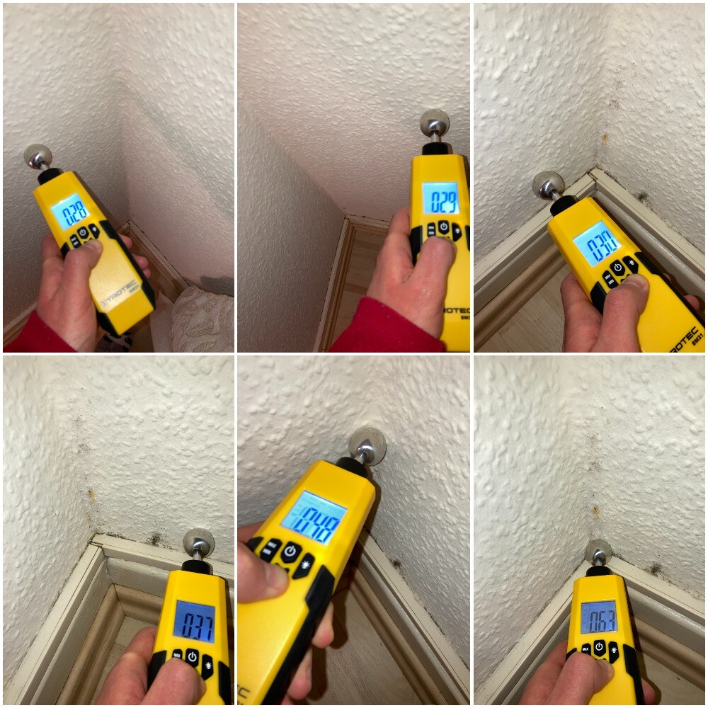 Yellow moisture meter held by hand checking corner of wall above baseboard; display shows values.