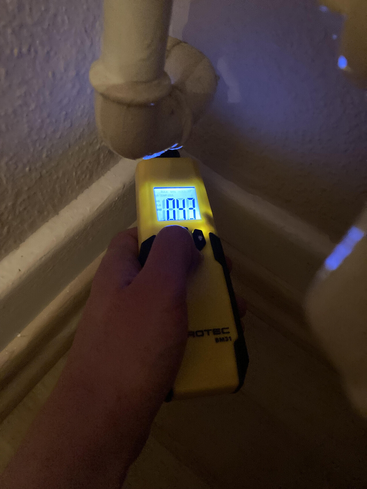 Person holding yellow meter under pipe connection; display lit blue showing reading.