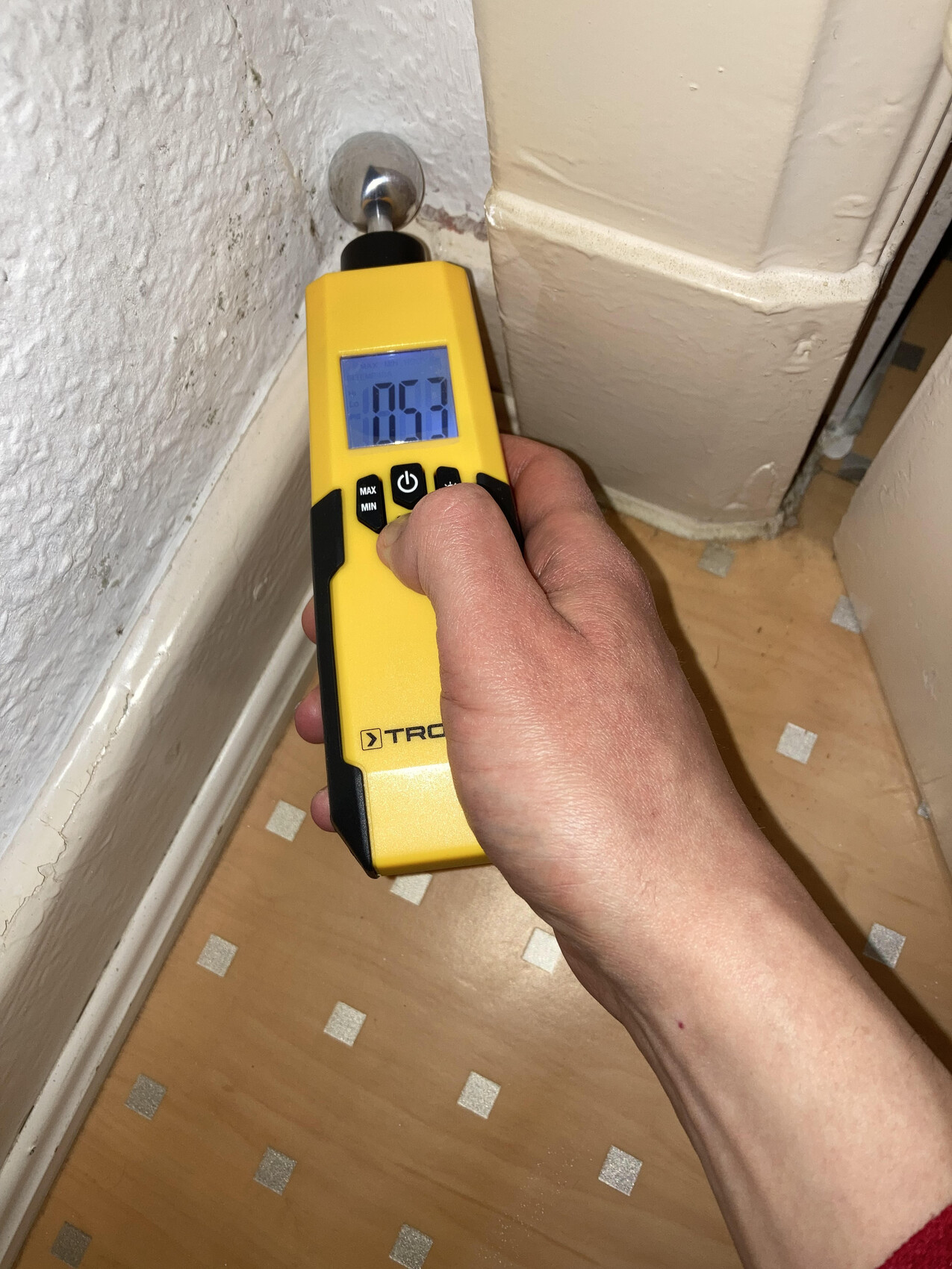 Yellow meter with display (0.53) held near wall corner by a pipe.