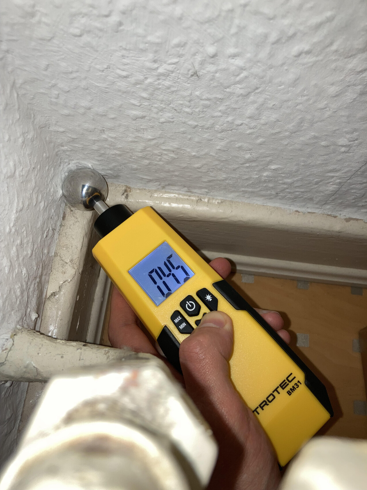 Yellow Trotec BM31 moisture meter with probes on window frame, reading visible.