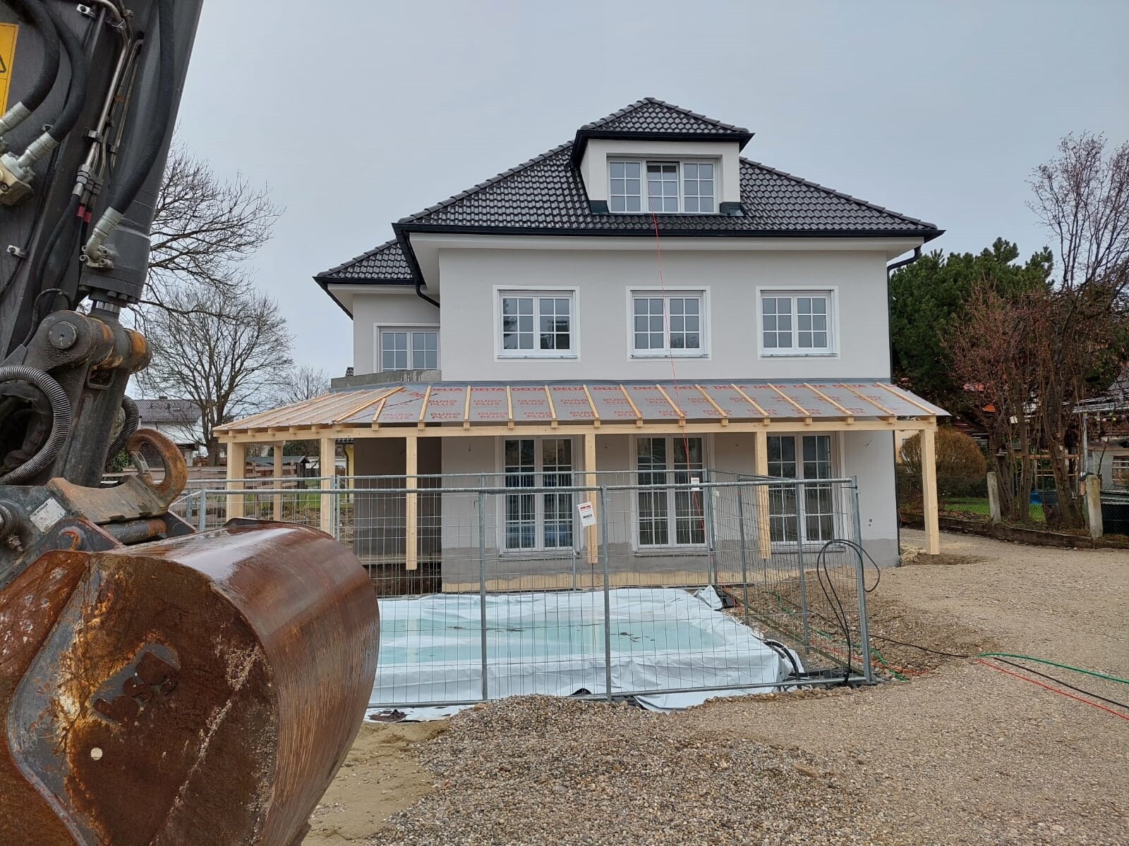 Mehrstöckiges Neubauhaus mit Veranda-Konstruktion, Bauzaun und Baufahrzeug links