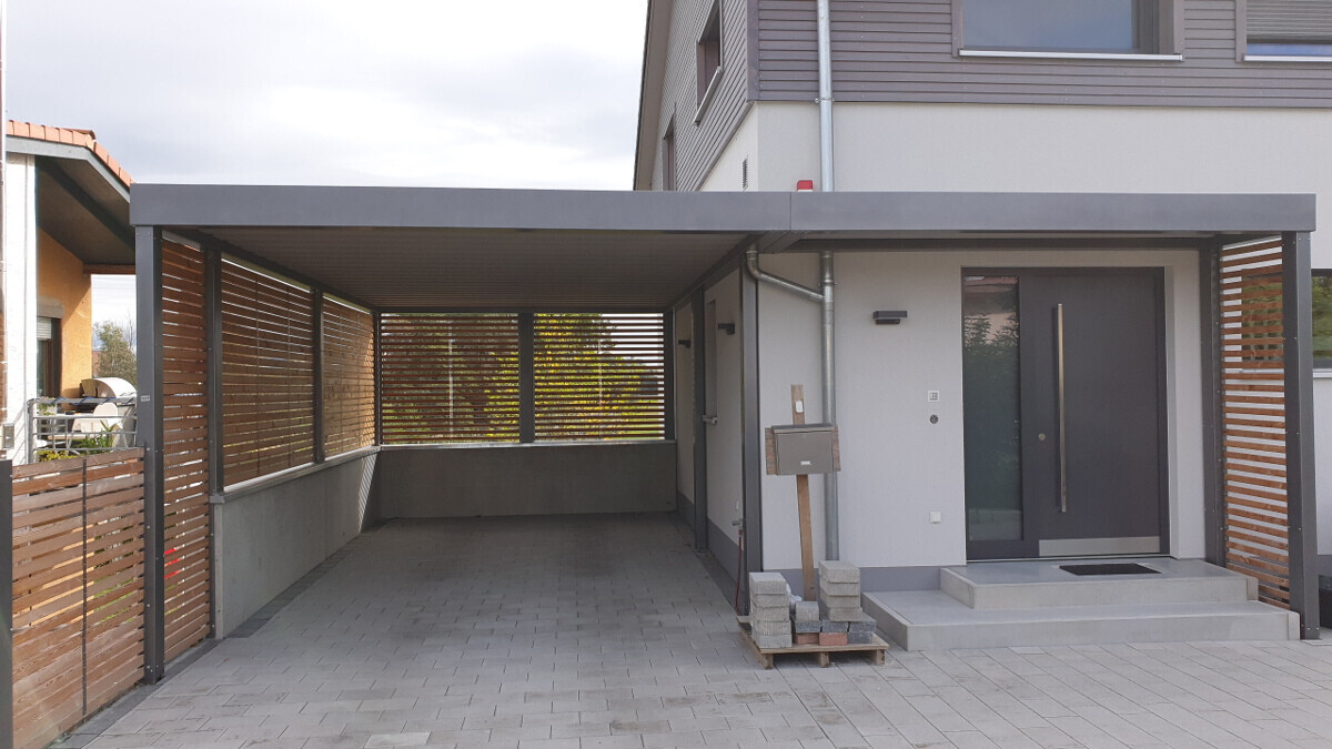 Modern driveway with carport, side wooden slats, gray entrance door, and mailbox.
