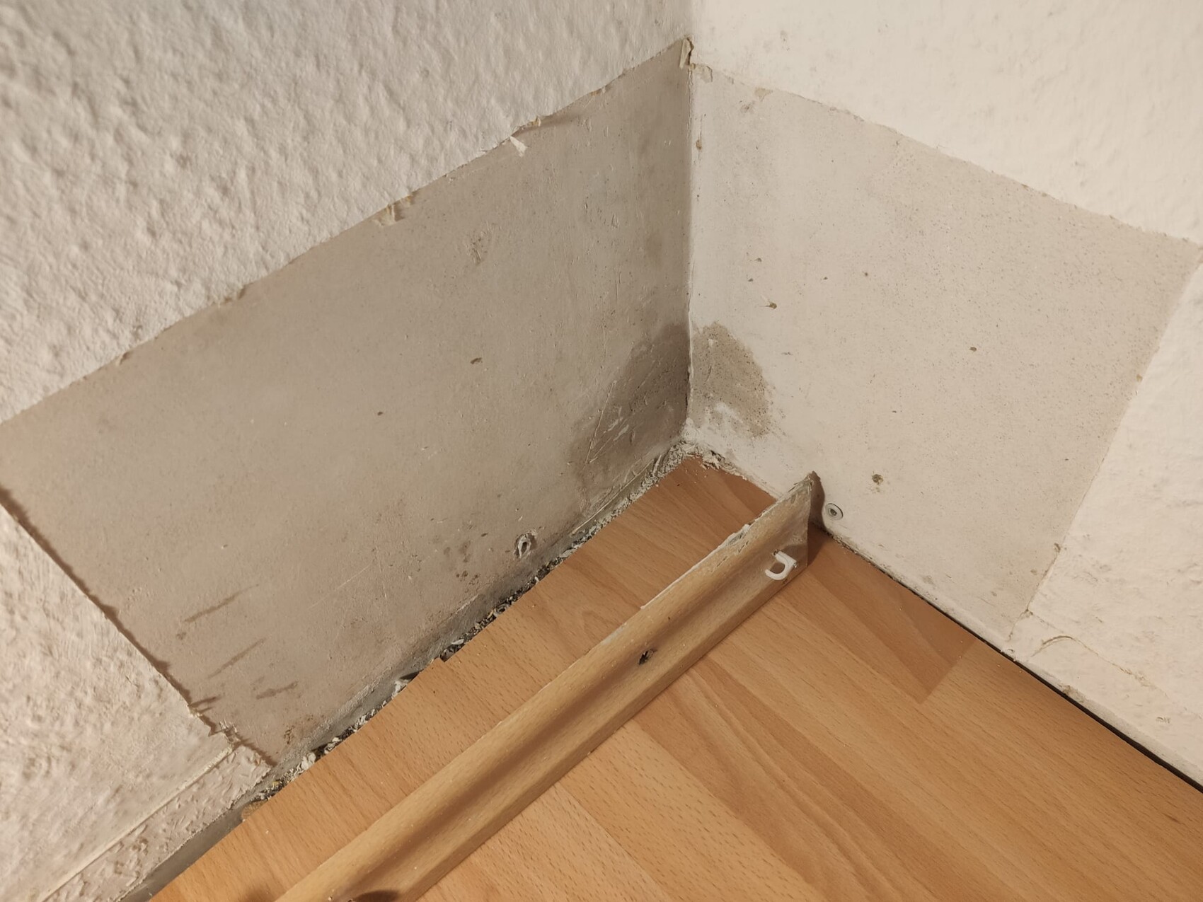 Corner of a room with damaged wall, plaster cracks, laminate flooring, loose baseboard.