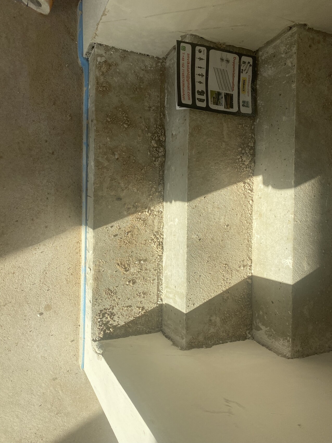 Baustellen-Ecke mit rohem Betonboden, kantigen Stützwänden und Schatten; ein Werbeblatt lehnt an der Wand.