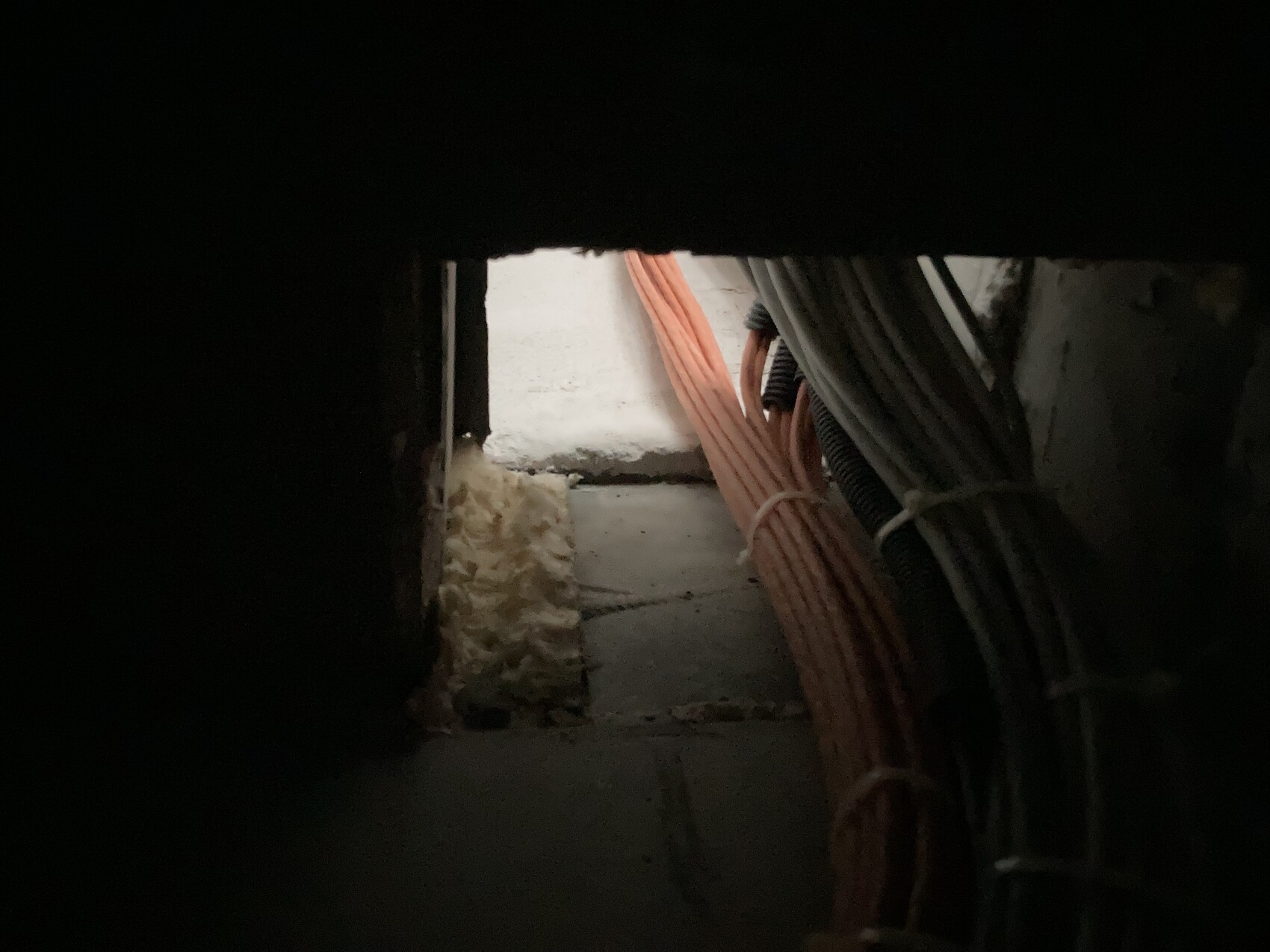 Dunkle Sicht durch Öffnung: freiliegte orange und graue Kabelbündel in einem Kellerraum.