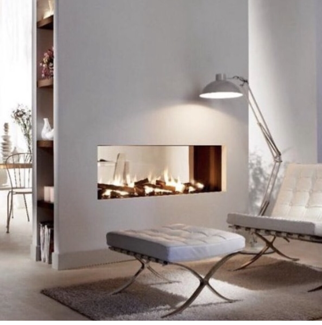 Modern living space with a burning wall-mounted fireplace, white lounge chairs, footstool, and reading lamp.