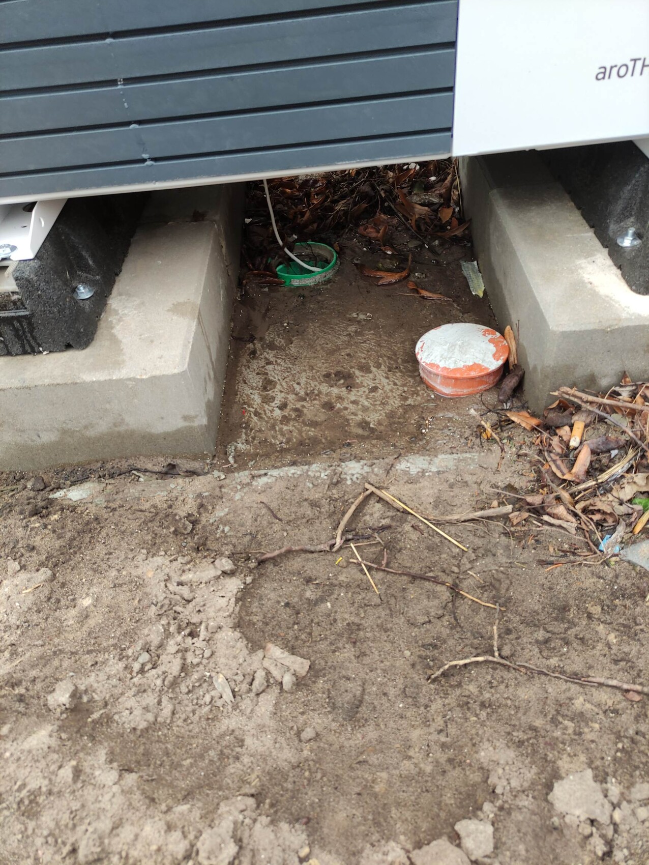 Outdoor unit on concrete blocks, open soil gap, green pipe cap and orange cover visible.