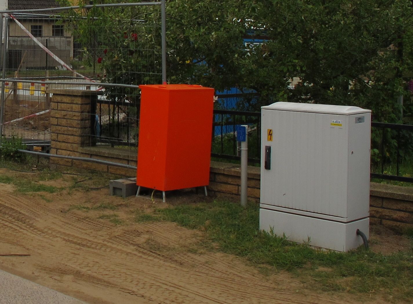 Orange metal box next to white electrical distribution panel at a construction site with fences.
