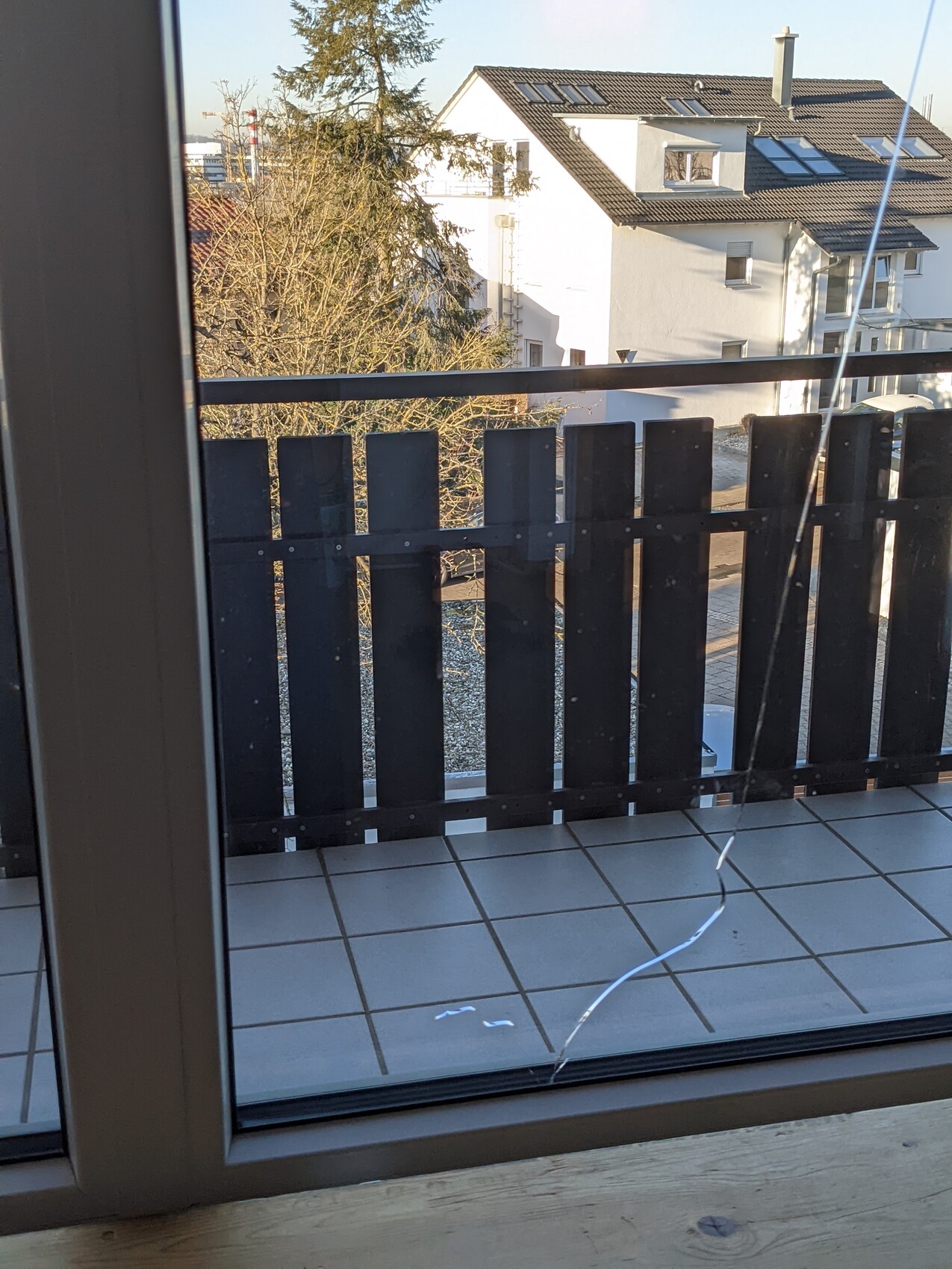 View through glass window onto balcony with dark wooden railing; houses, tree, tiled floor.