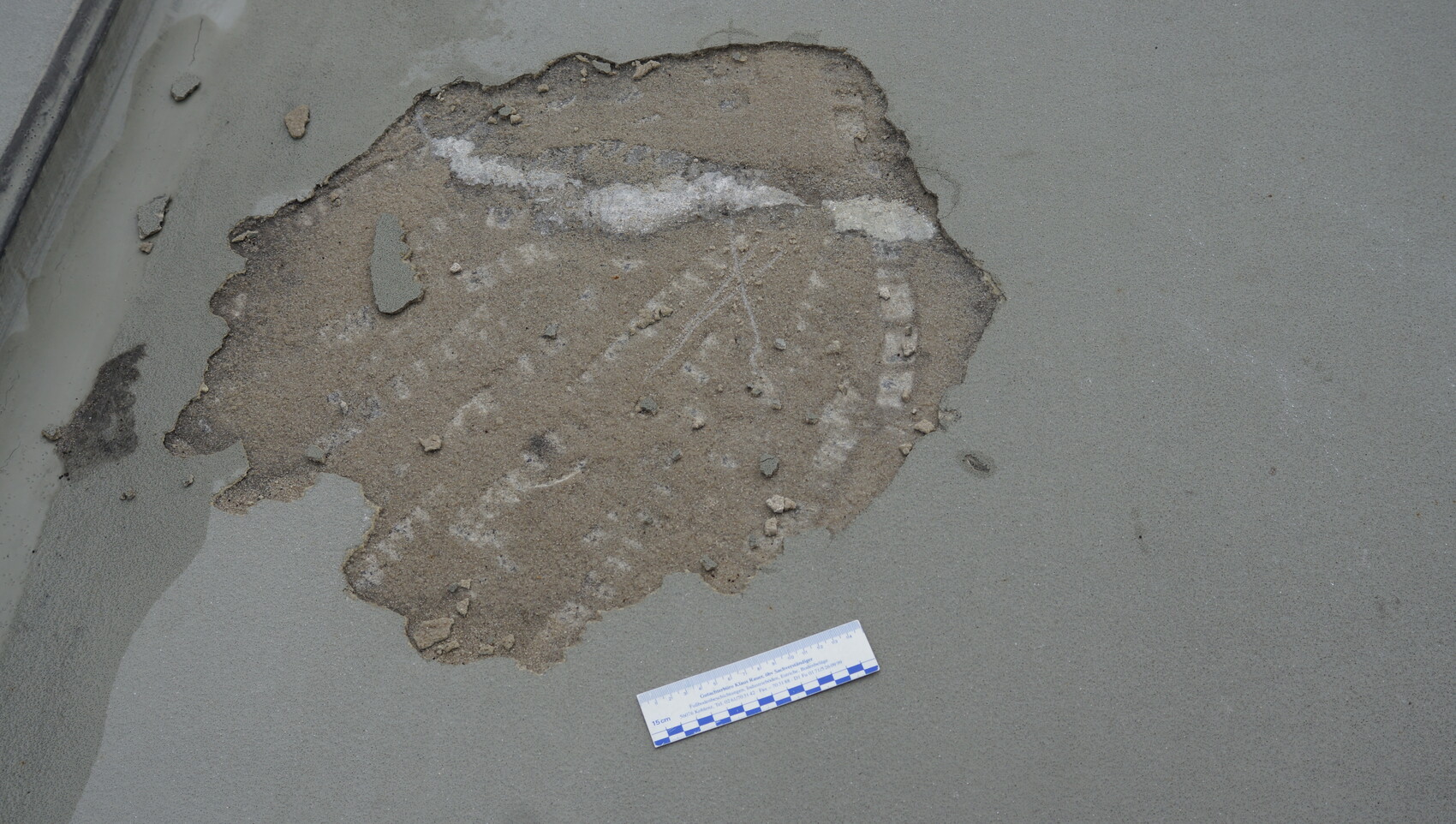 Partially exposed soil and gravel layer in a damaged concrete surface, adjacent to a measuring scale.