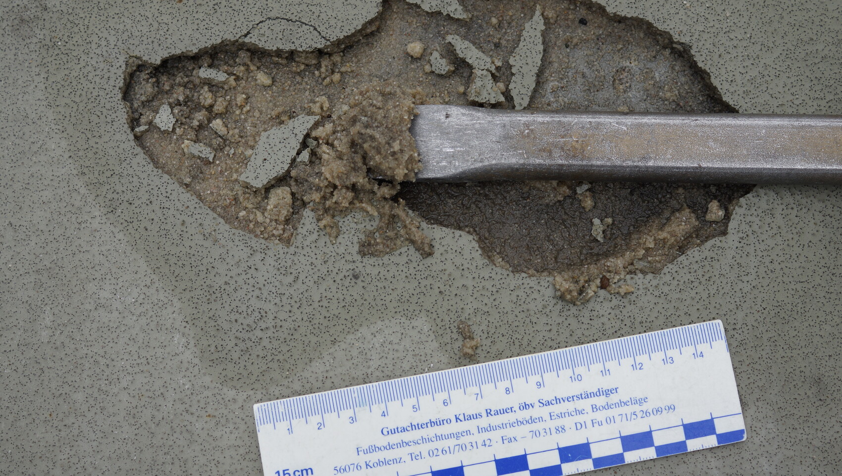 Cracked concrete floor with an open fracture; a metal bar runs over it; measuring rod below for scale.