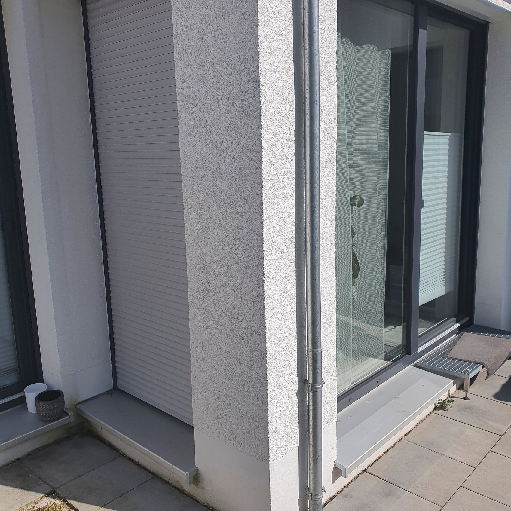 White facade with downspout, large glass front with roller shutters and terrace.