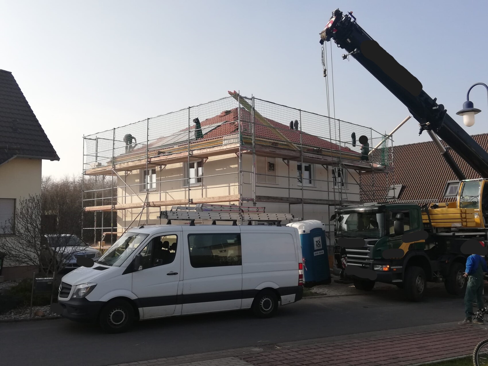 Construction site: single-family house with scaffolding, roof work; crane, white van and porta-potty on site.
