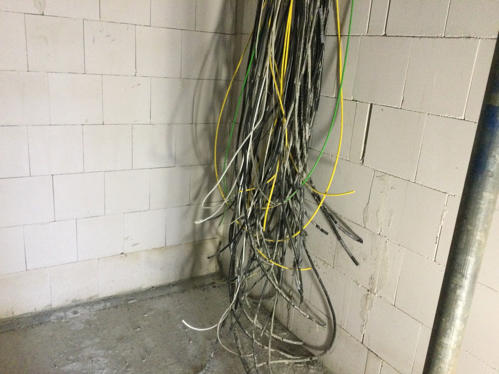 Loose bundles of black, yellow, and green wires hanging on a white concrete block wall.