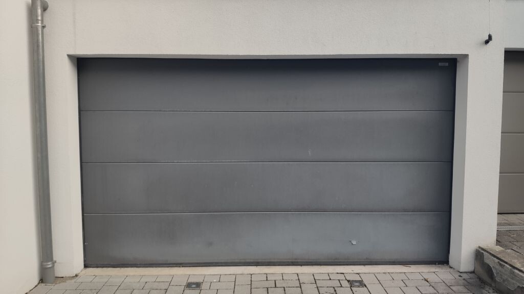 Closed gray metal garage door in front of a white house wall, with a downpipe visible on the side.