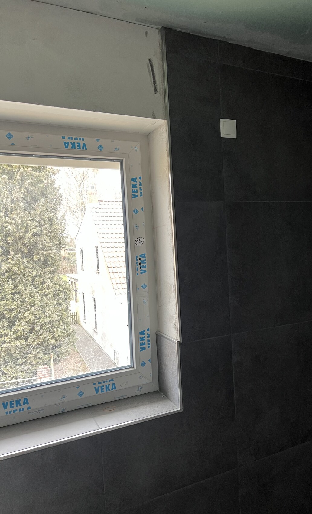 New window with VEKA protective film in a dark tiled wall; view outside of trees and roofs.