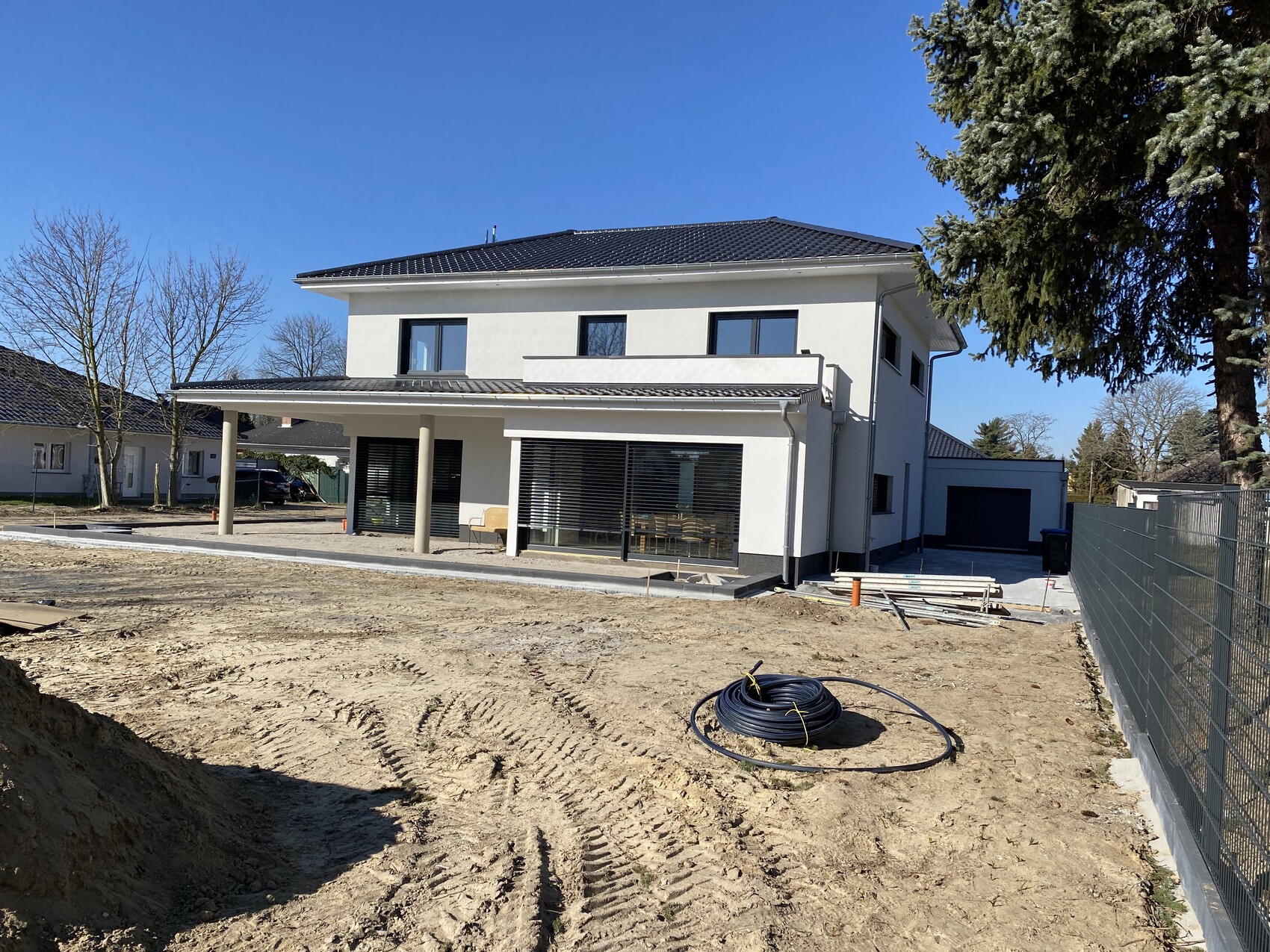 Modern two-story detached house with terrace; building site in front, cable drum and building materials.