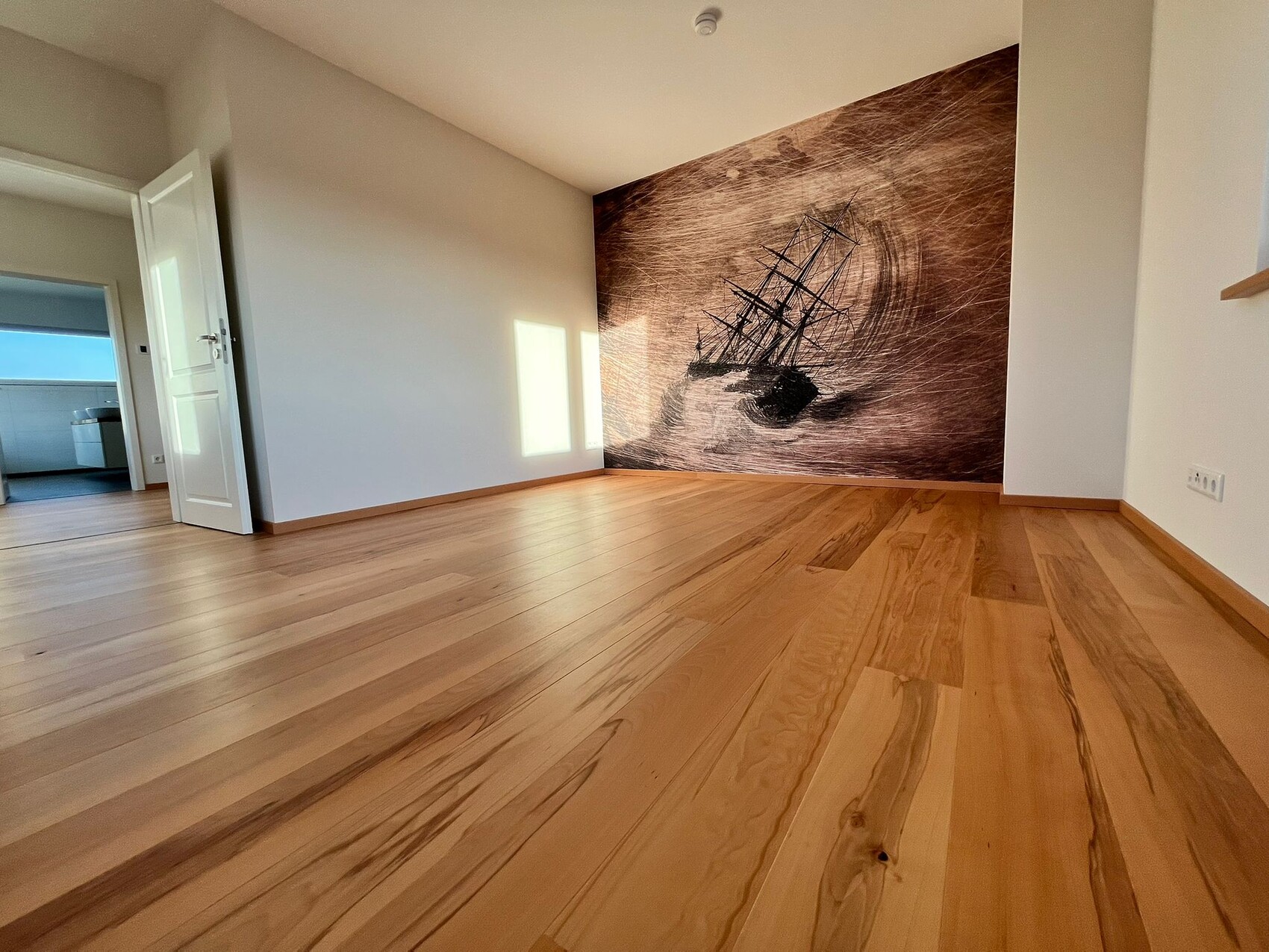Empty living room with light wooden floor; wall painting of a sailing ship on the back wall.