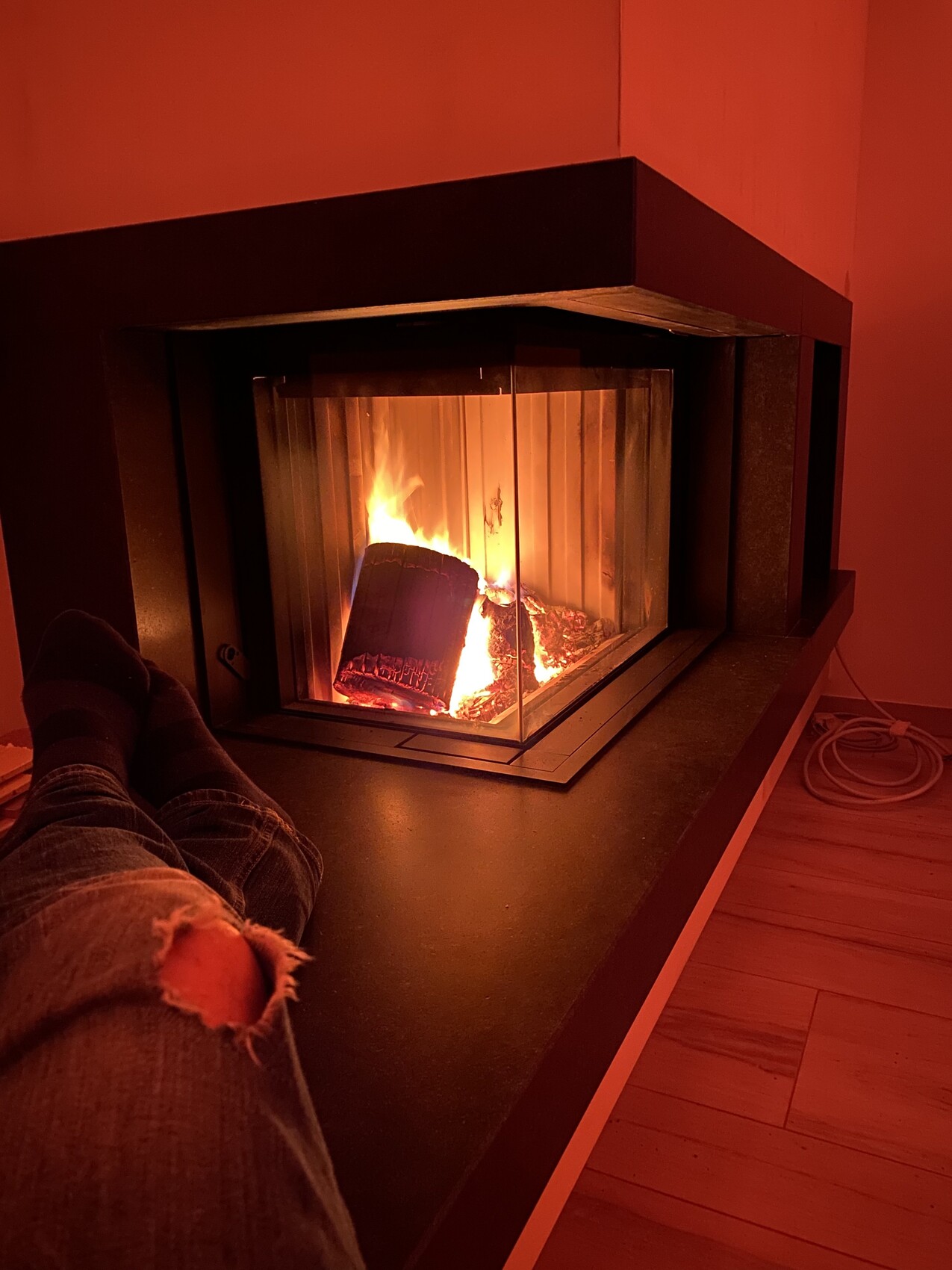Person's legs in front of a fireplace with burning fire, red lighting; jeans with a hole.