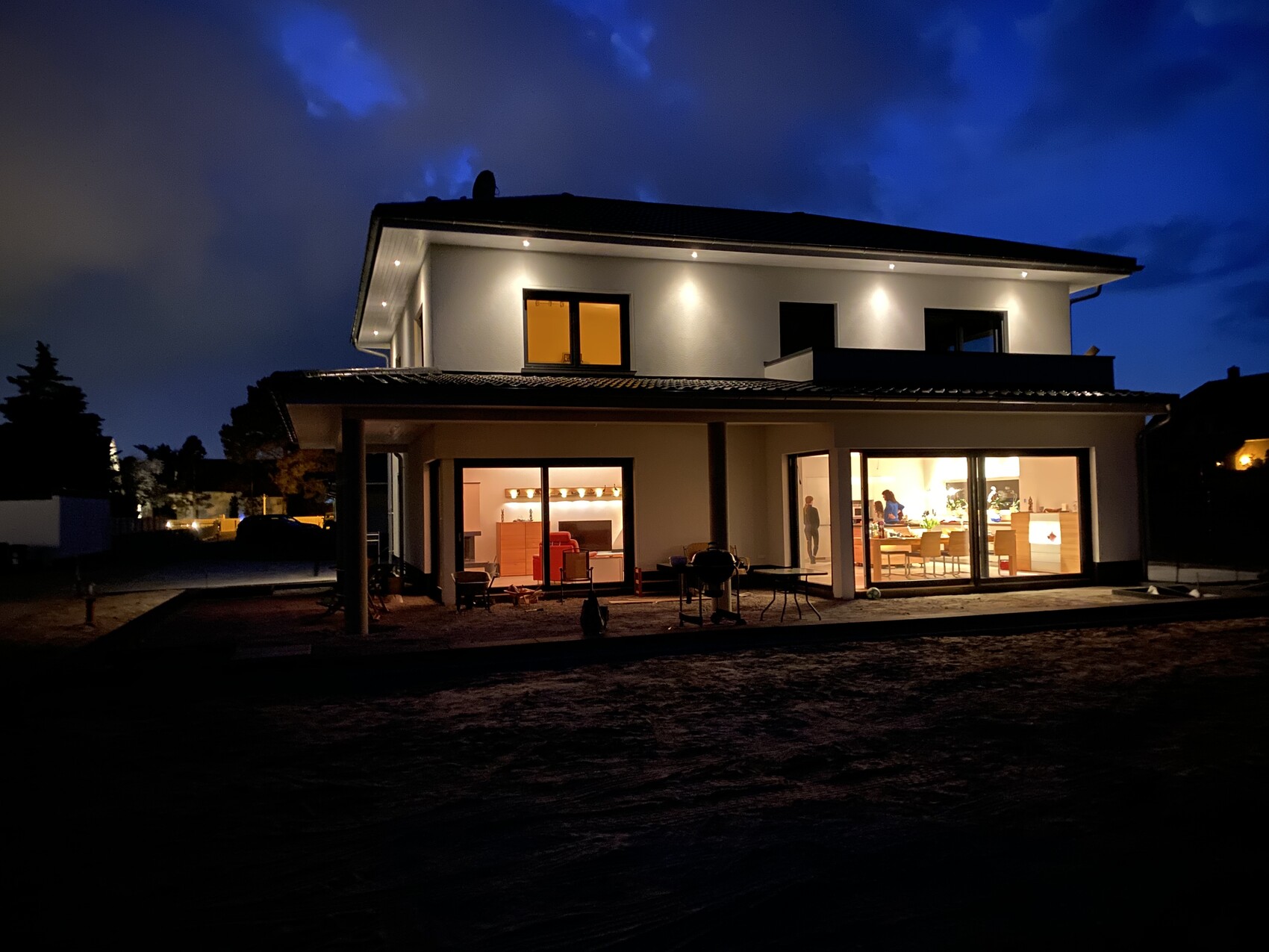 Modern two-story house at night, bright interior with living room and kitchen visible through glass windows.