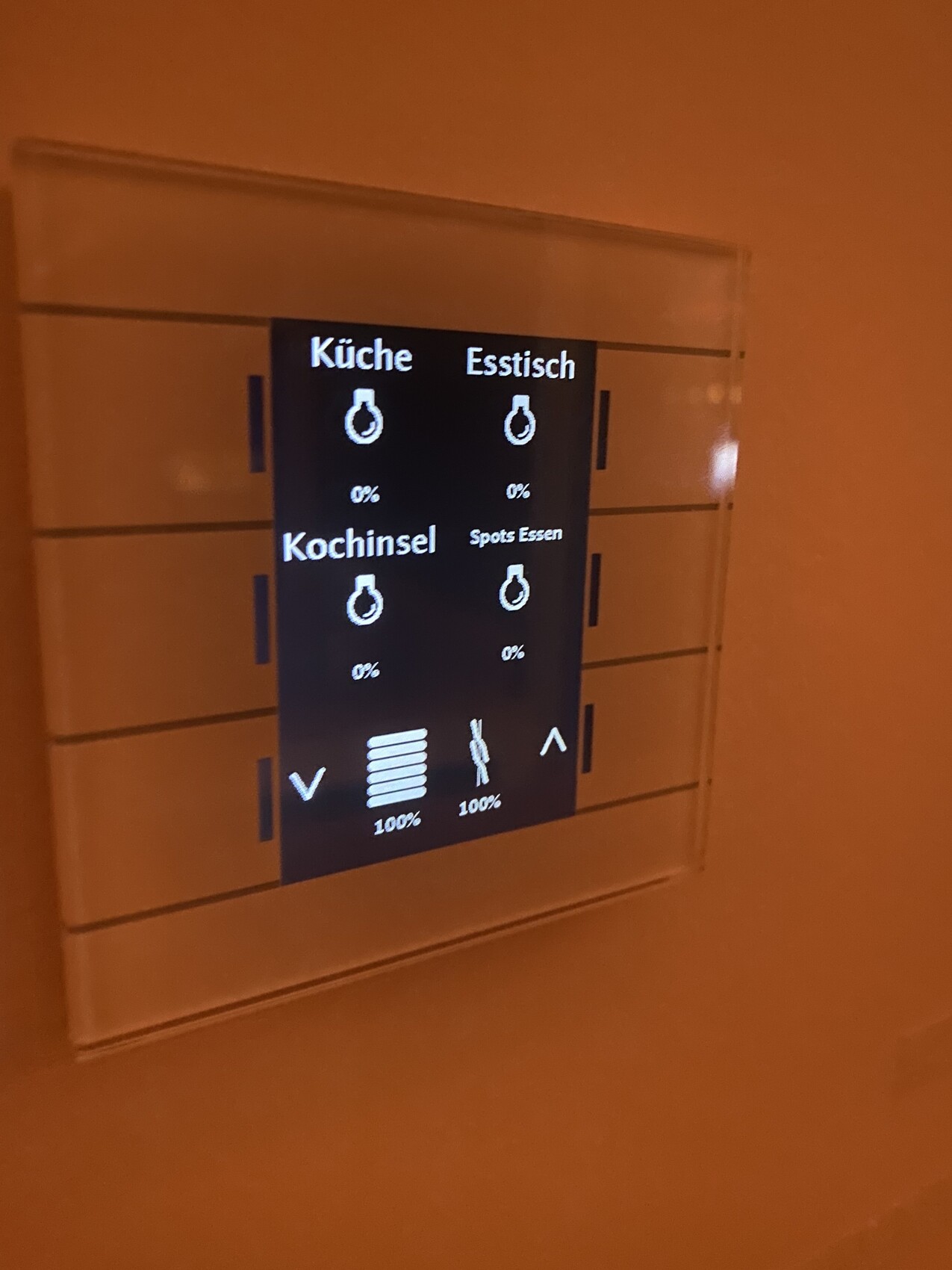 Smart home touchscreen in a wall niche, showing kitchen, dining table, kitchen island, and spotlights.