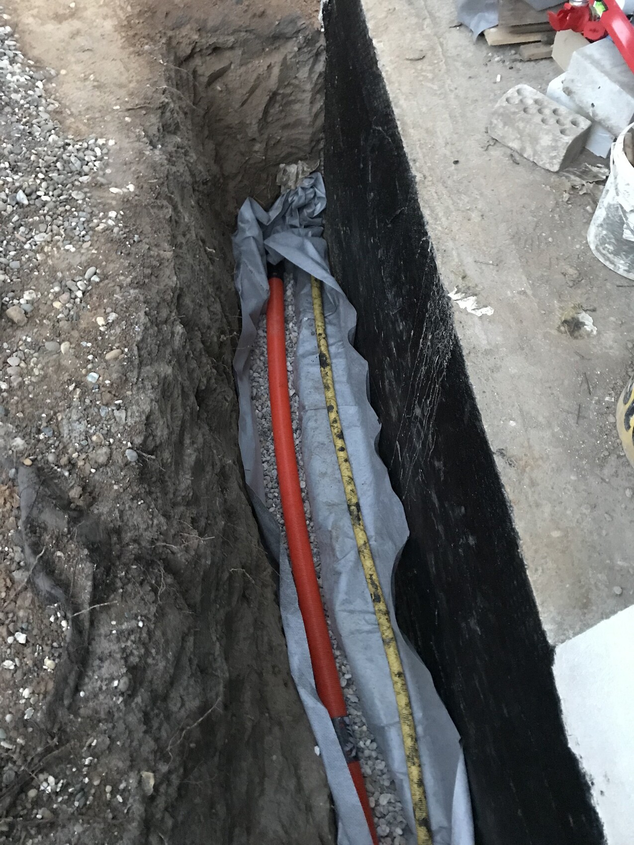 Construction pit with red pipe, yellow tape measure, and gray protective foil next to black wall