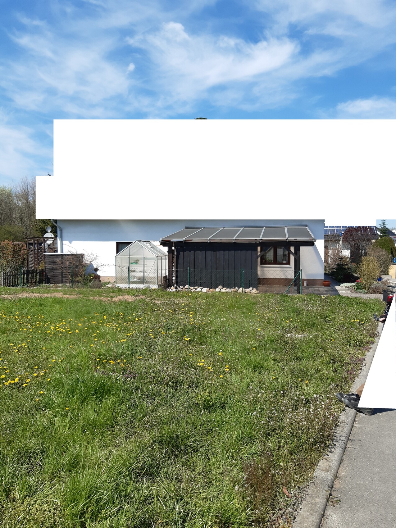 Front view of a single-family house with greenhouse, carport and lawn with dandelions.