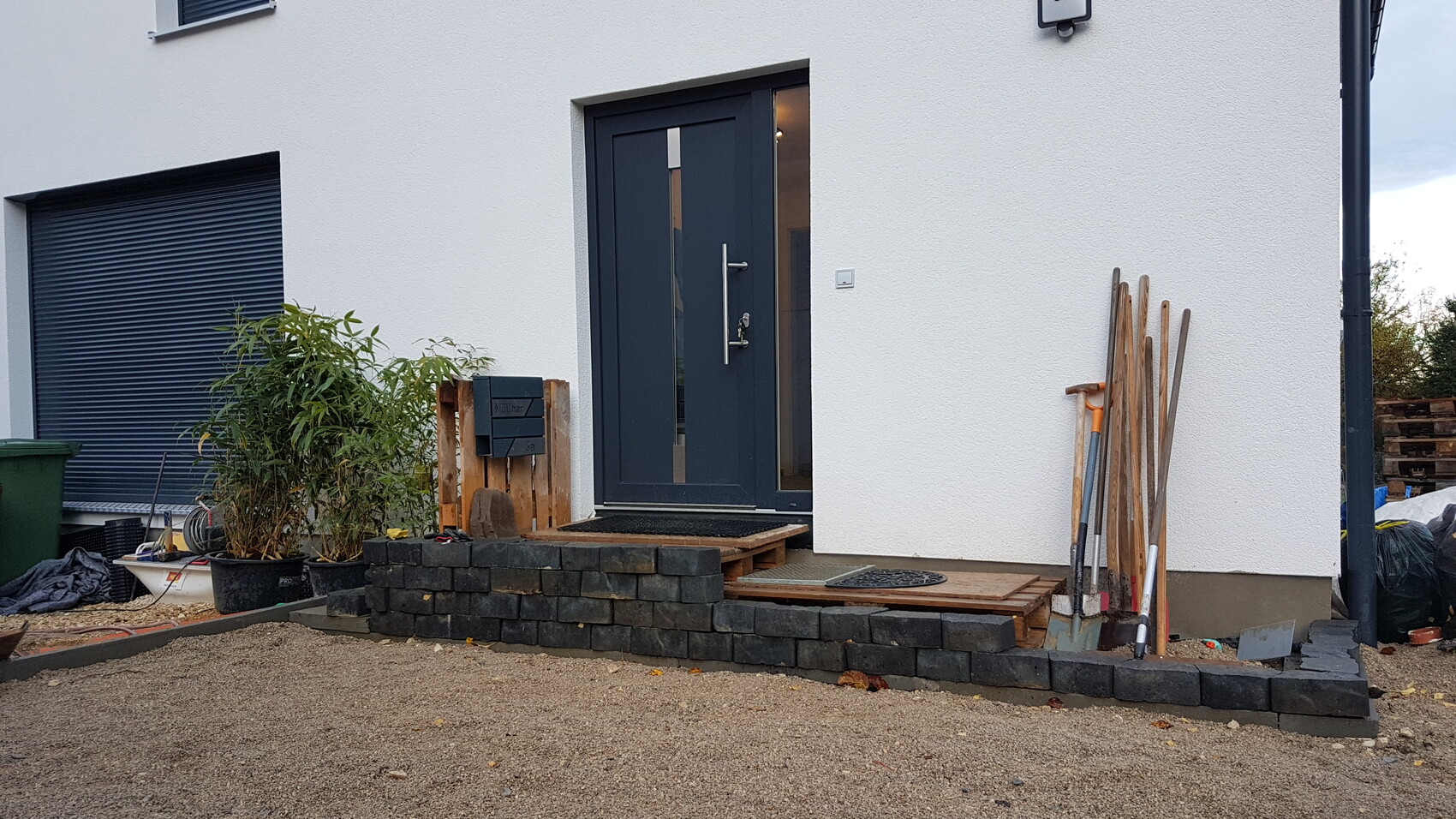 Modern front door in dark gray in front of white facade; building materials, plant and tools at the entrance.