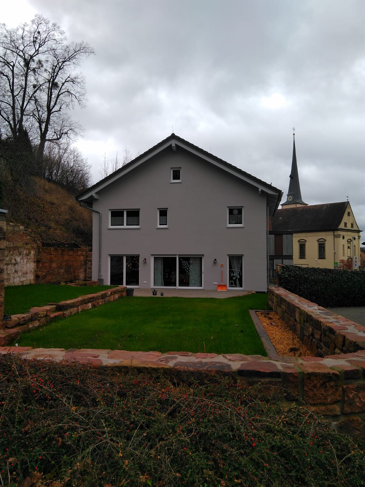 Gray two-story house with garden, church tower in the background, cloudy sky.