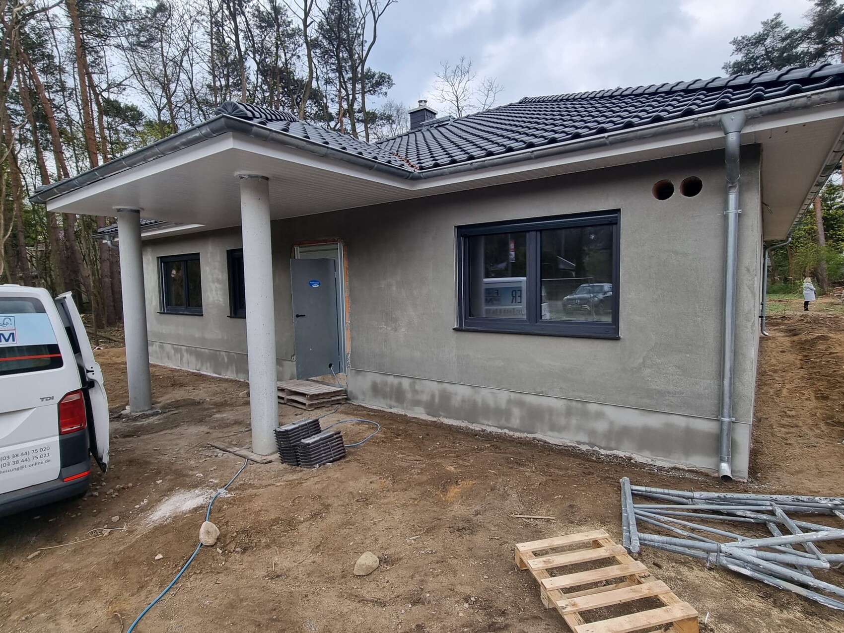 New single-family house under construction with covered front terrace, construction materials in front of forest.