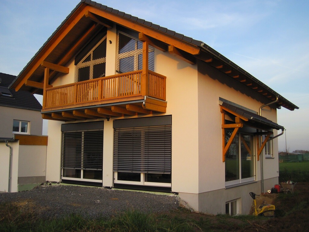 Two-story house with wooden balcony, large windows and roller shutters, front garden.