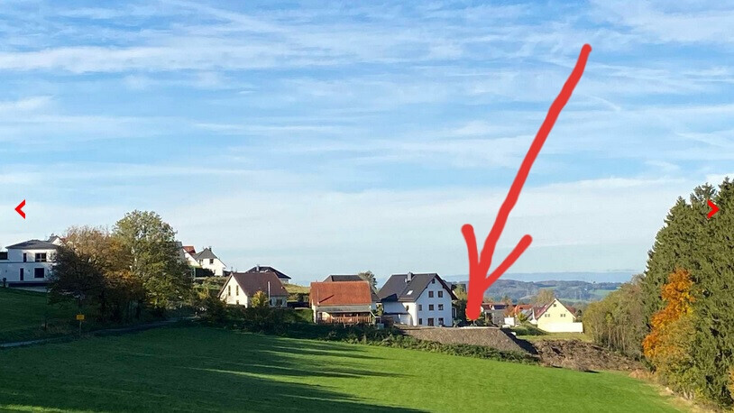 Small village with white houses on a hillside, green meadows in front; red arrow pointing to one house.