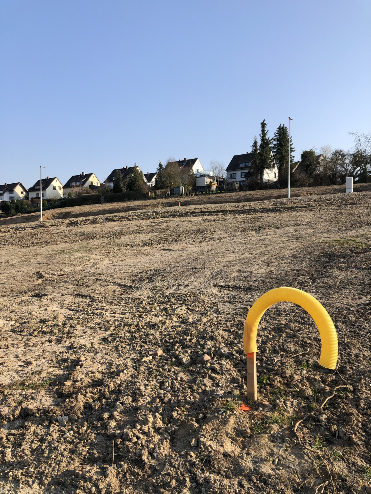 Sparse construction site with yellow curved pipe stub in sand, houses in the background.