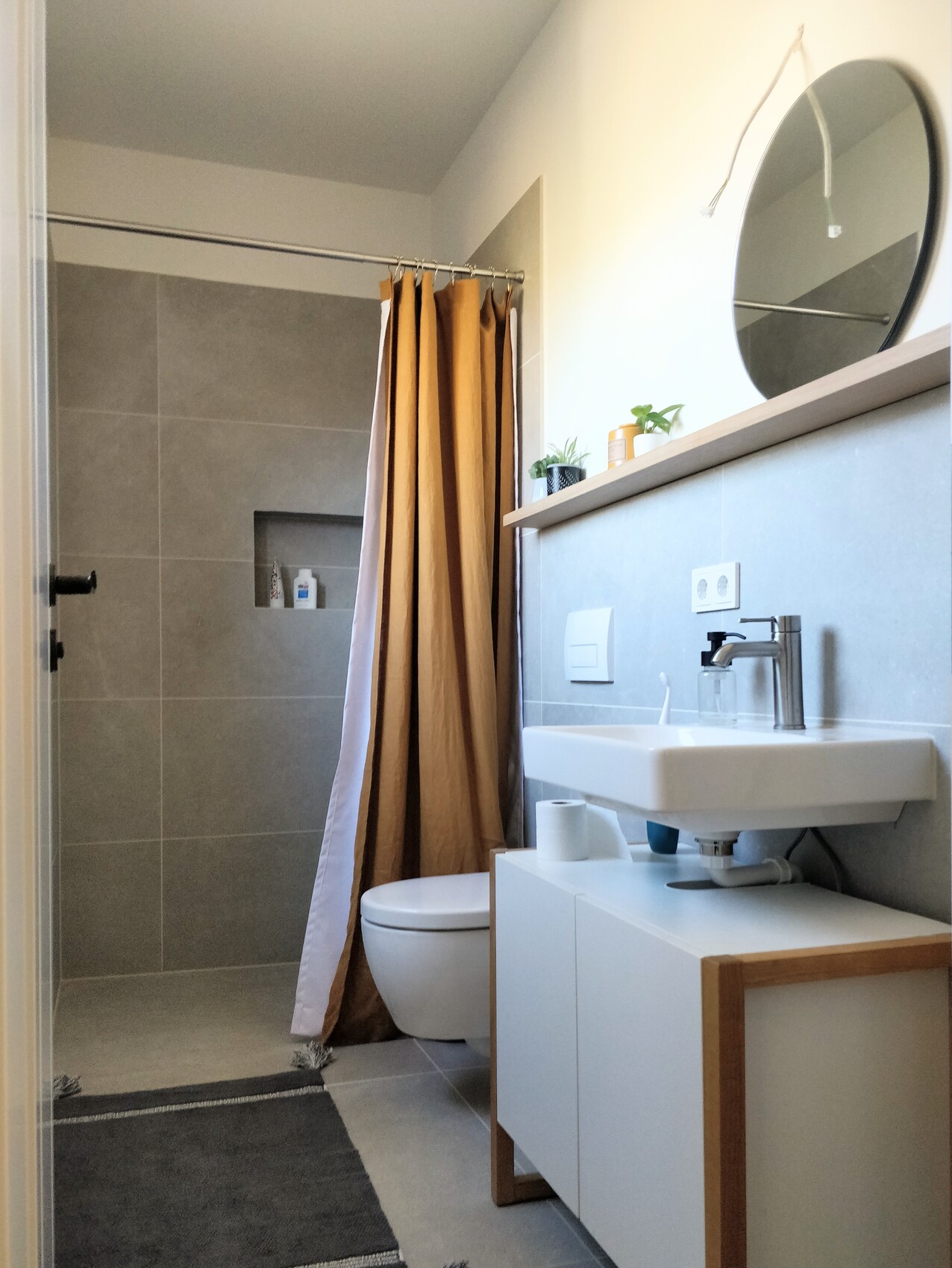 Bathroom: shower with brown curtain, toilet, sink, round mirror, plants.