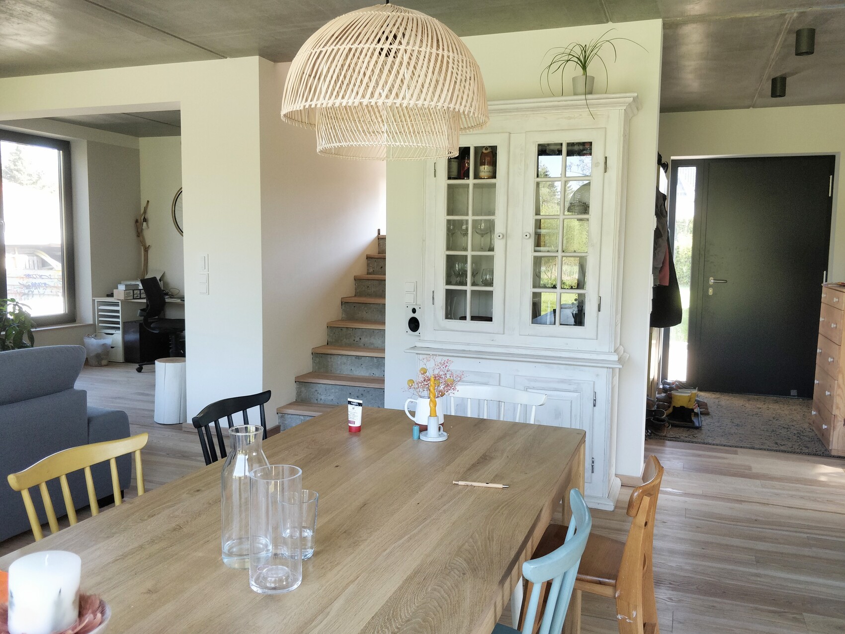 Dining room with wooden table, glass cabinet, colorful chairs, rattan pendant lamp, staircase in the background.
