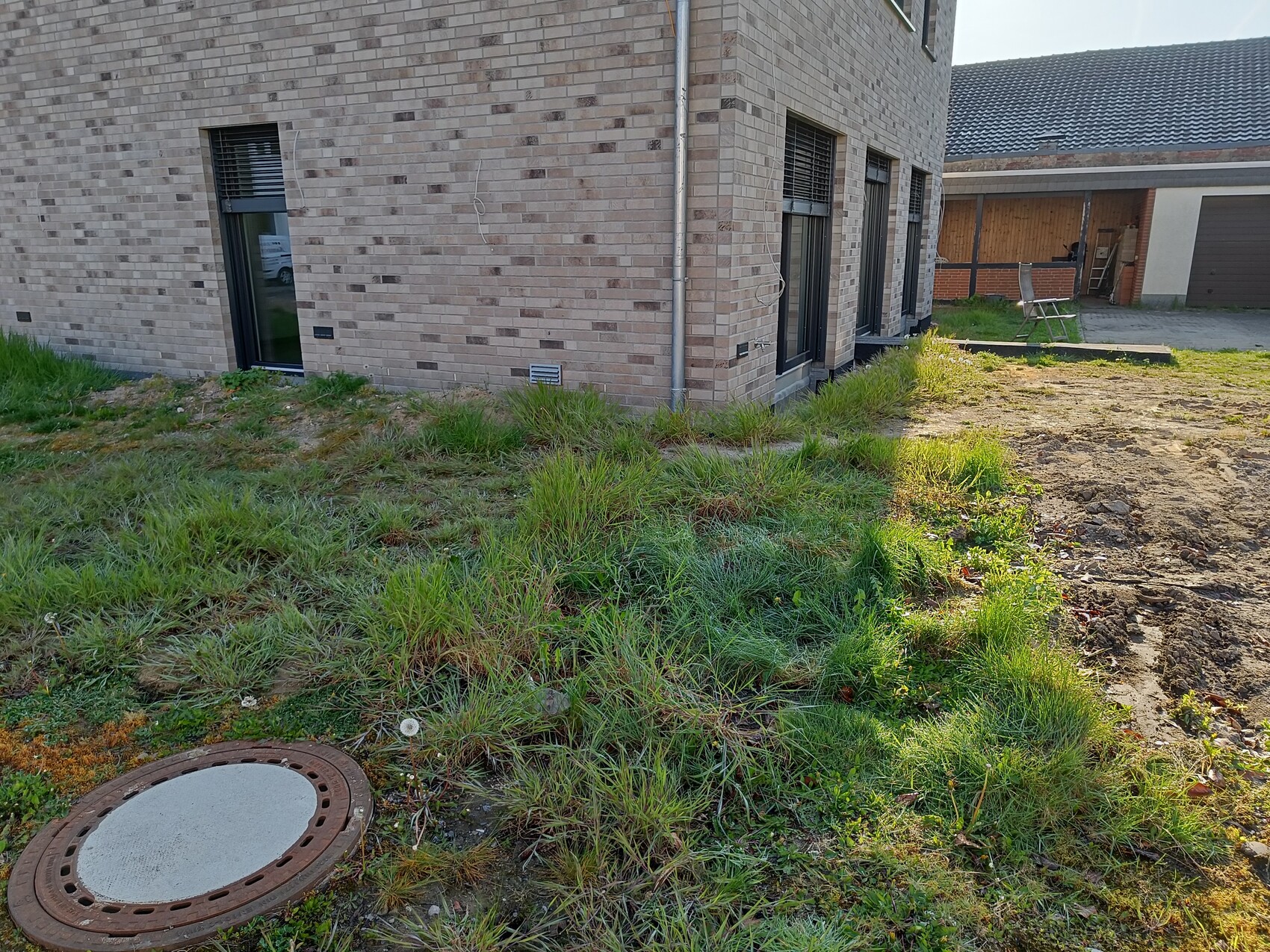 Außenansicht eines Backsteinhauses mit ungemähtem Gras und runder Schachtdeckel im Vordergrund.