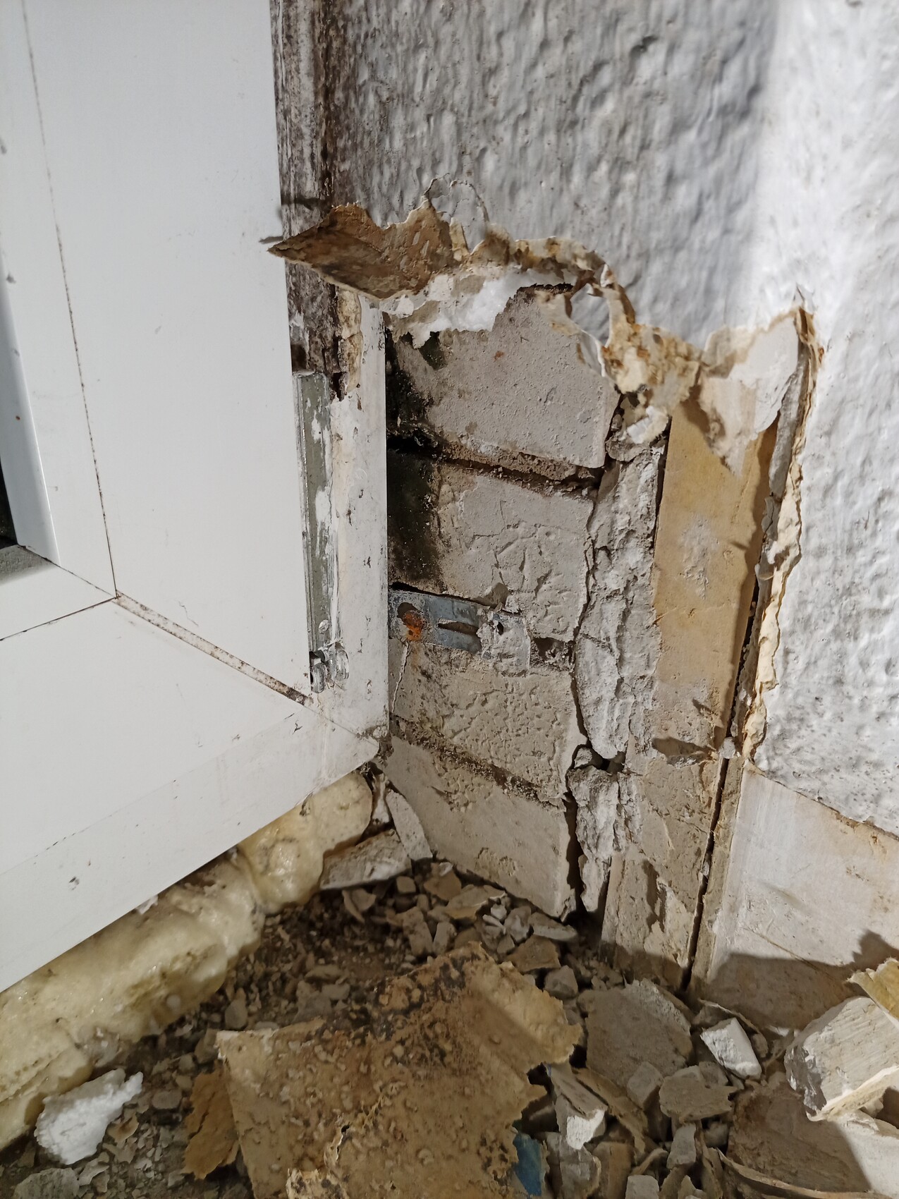 Damaged interior wall: exposed concrete block, plaster break, debris on floor next to window frame
