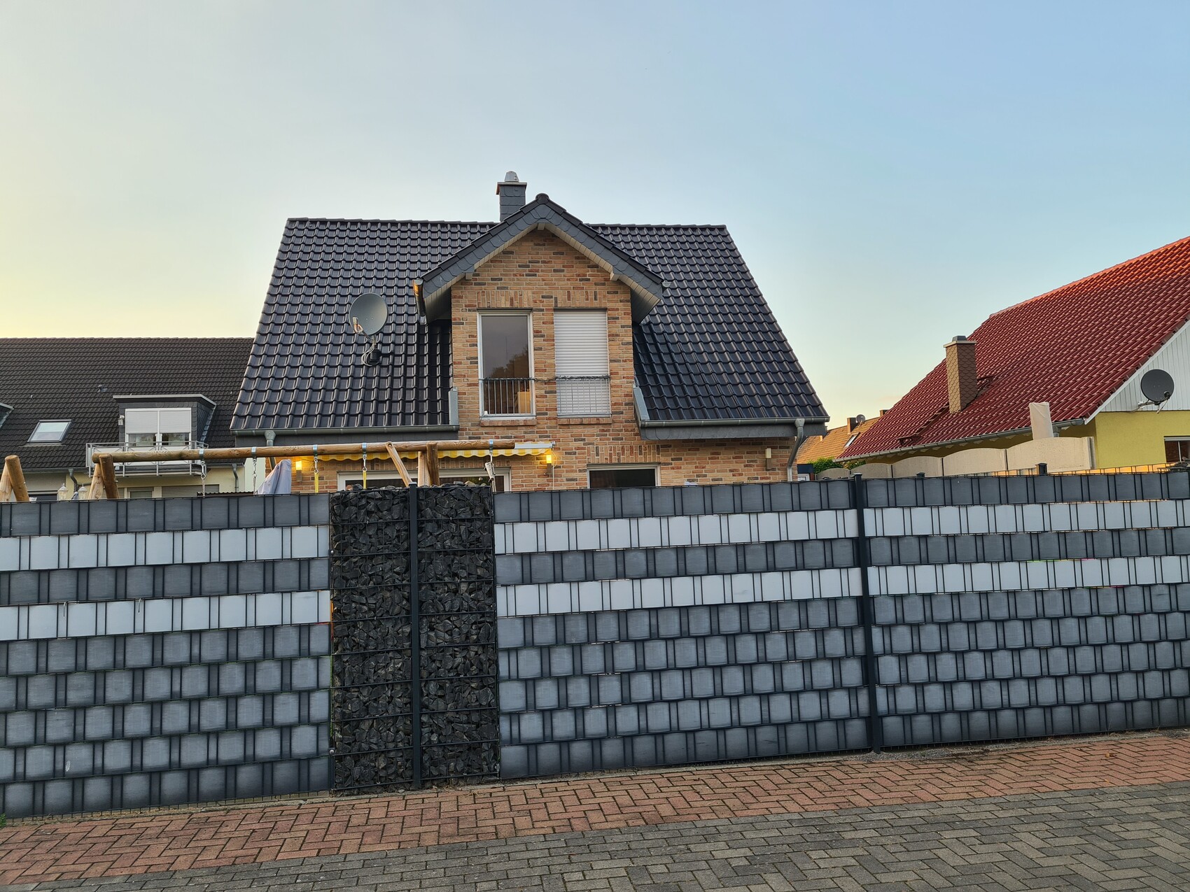 Two-story brick house with dark tiled roof behind a gray panel fence and gabion pillar.