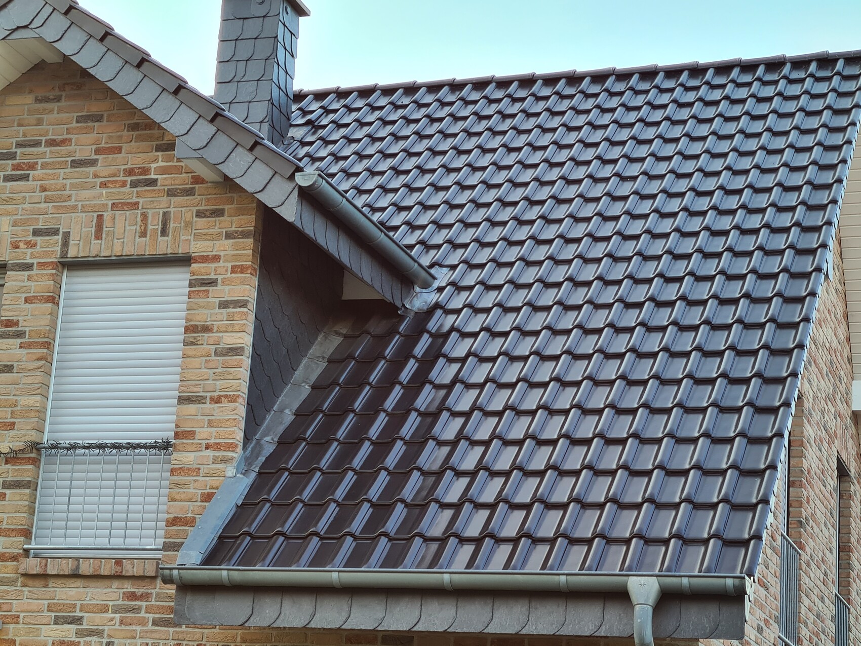 Roof of a brick house with brown roof tiles, chimney and roller shutter windows.