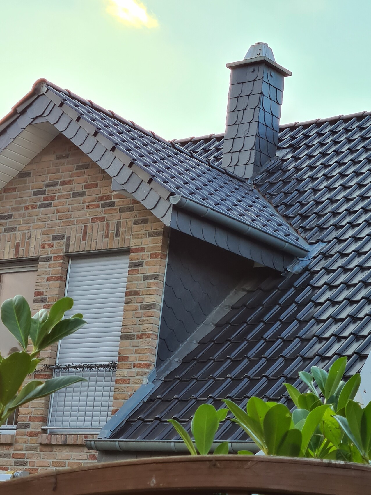 Roof of a house with gray slate tiles, chimney, window, green leaves in the foreground.