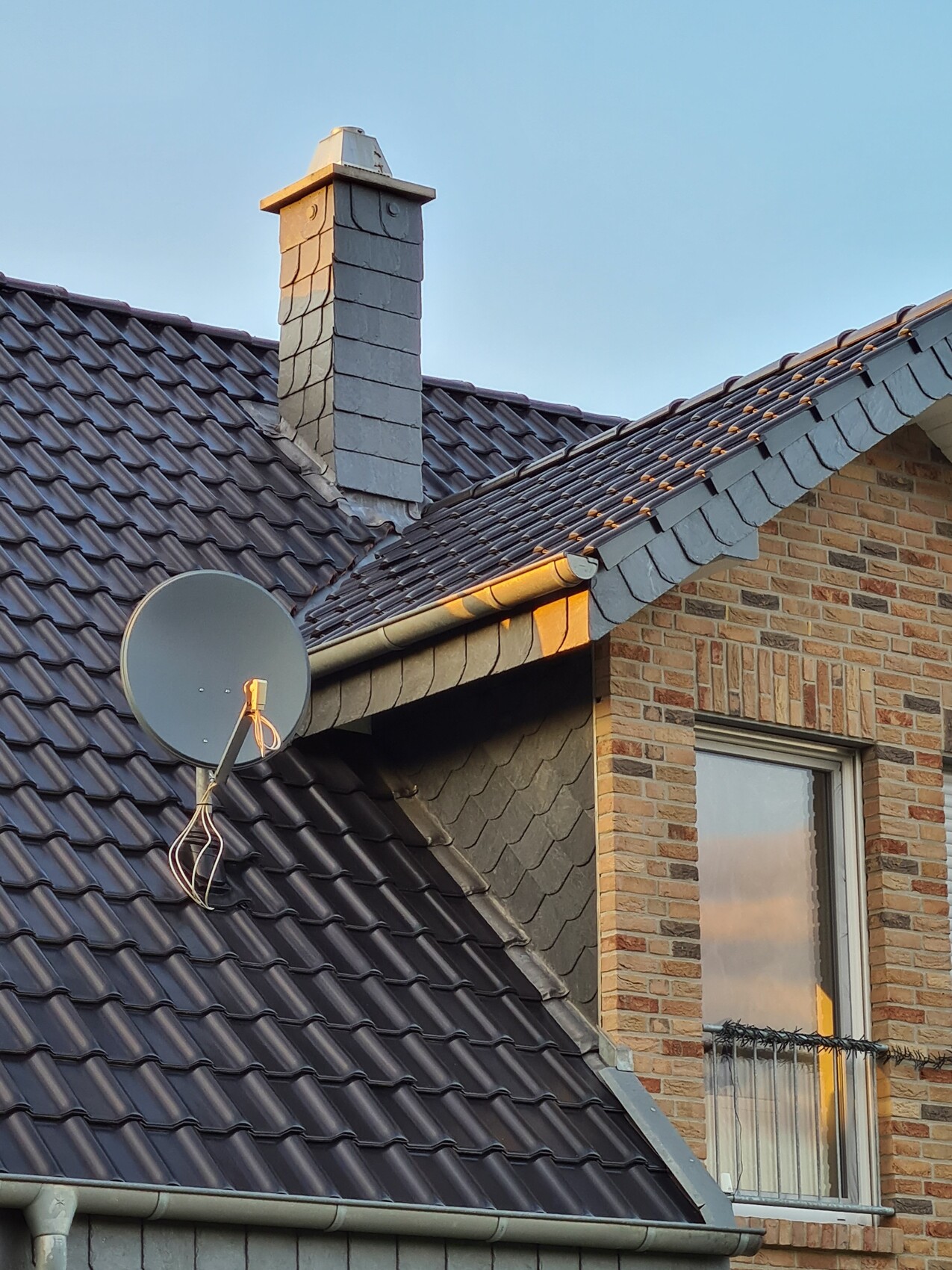 Roof of a house with gray tiles, chimney, satellite dish and window.