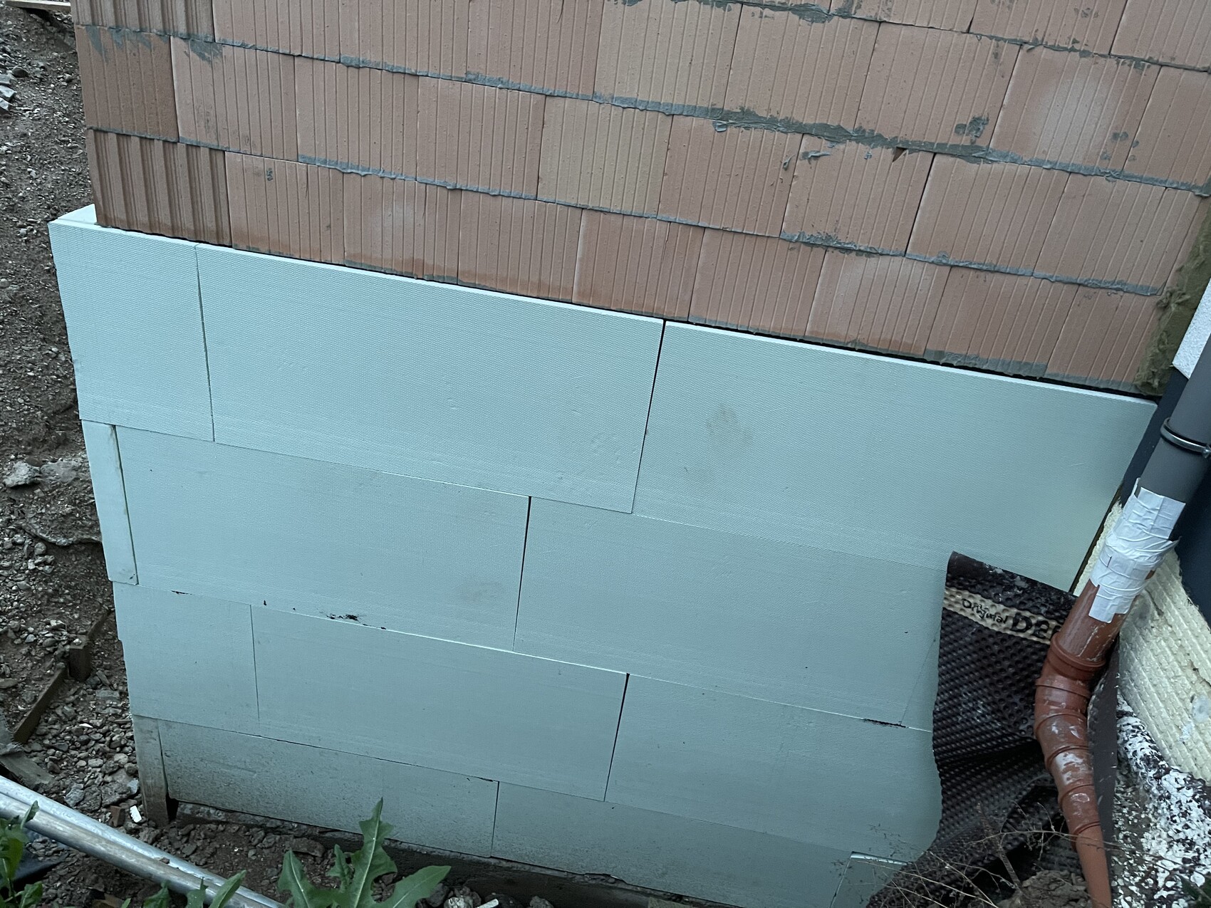 Exterior wall with light blue insulation boards (polystyrene) beneath brick wall; piping at the edge.