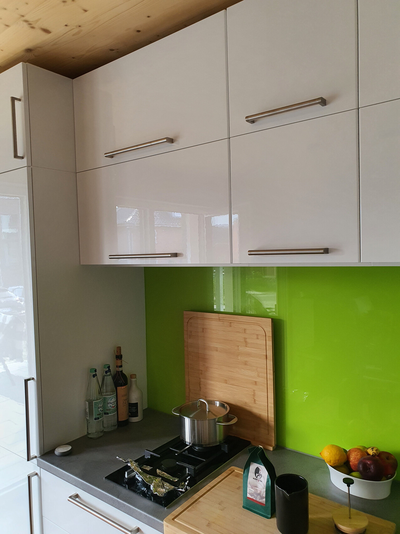 Kitchen with white high-gloss cabinets, green backsplash, pot on gas stove, fruit bowl.