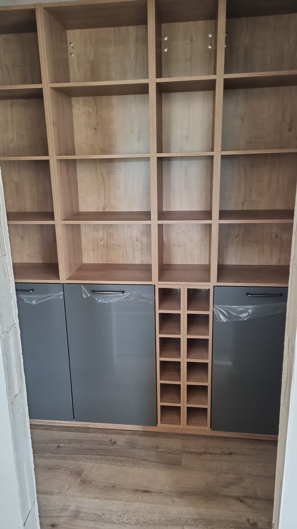 Large wooden shelf with many open compartments, small bottle holder in the middle, gray base cabinets.