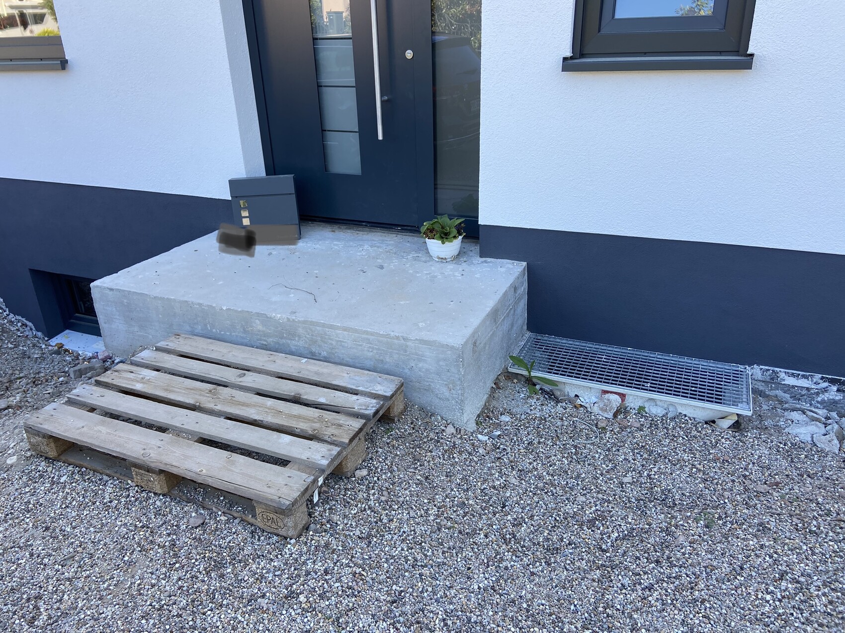 Exterior view of a house: dark front door, wide concrete step, potted plant, and gravel ground