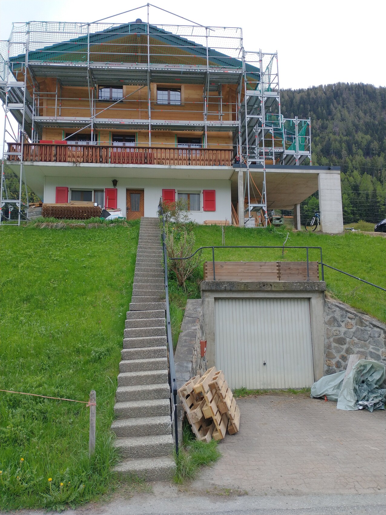 Multi-story house with scaffolding, green lawn in front, stairs leading to the entrance, garage below.