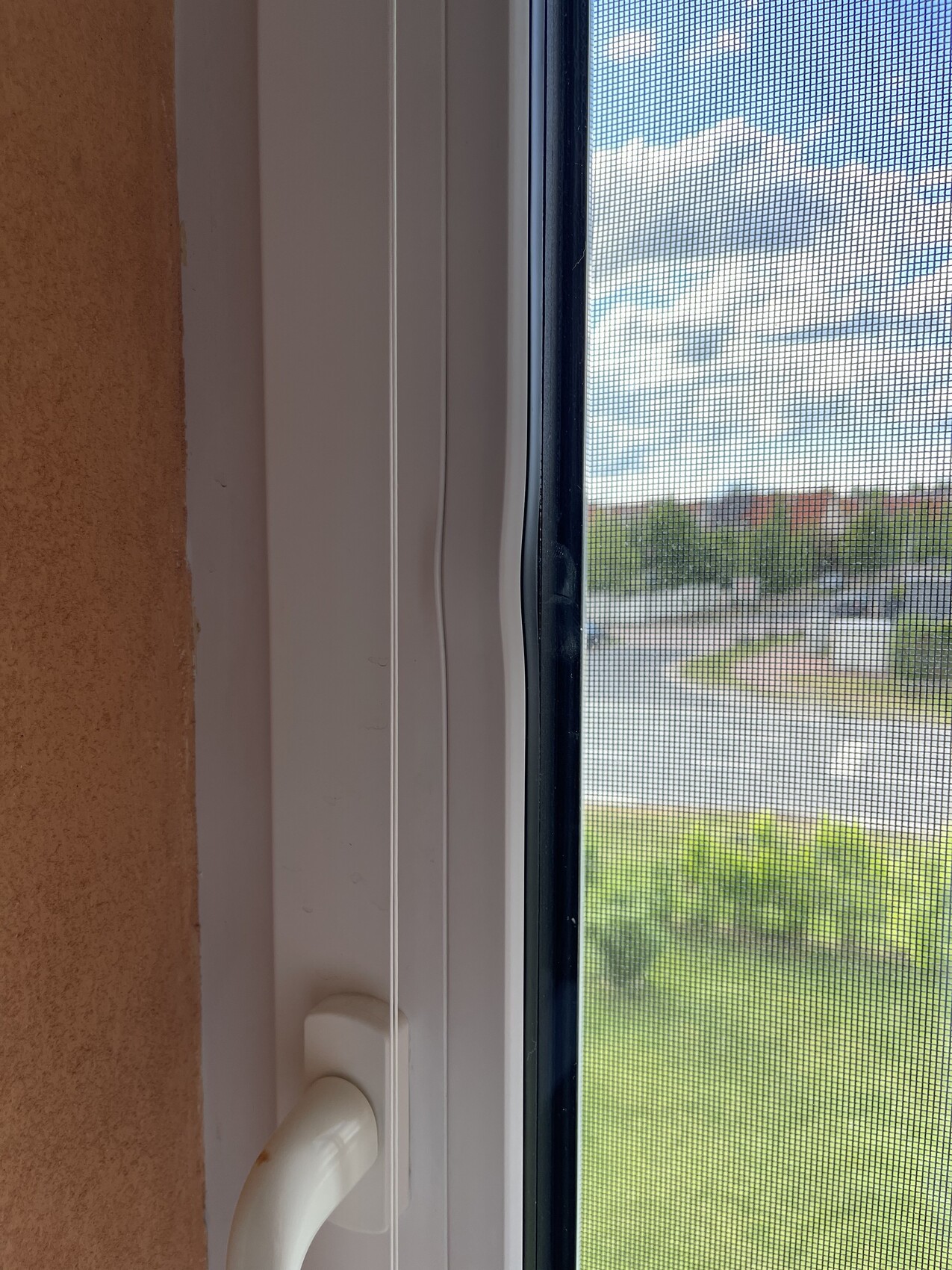 Window frame with insect screen, handle at the bottom; view outside to green meadow, road, and distant houses.