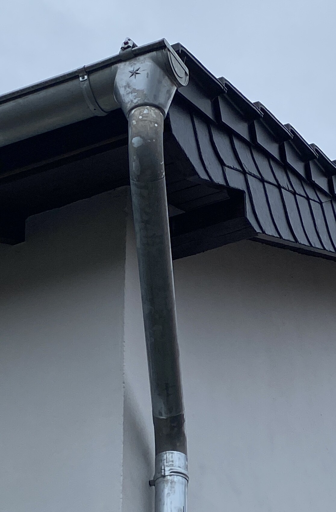 Vertical metal downspout of a rain gutter at the building edge under a dark tiled roof.